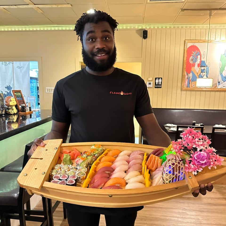 A smiling man in a Flame Sushi shirt holds a large wooden boat filled with various sushi and sashimi.