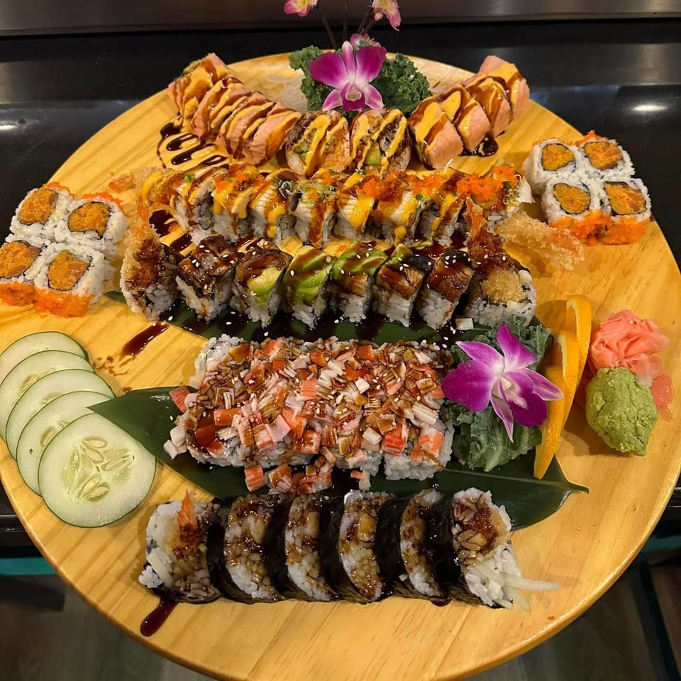 A large wooden platter overflowing with various Flame Sushi rolls, garnished with orchids and cucumber slices.