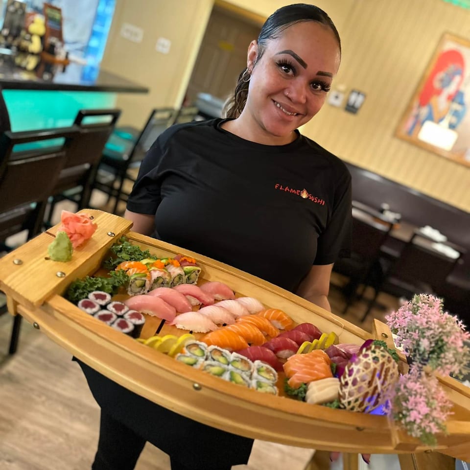A smiling Flame Sushi server holds a large wooden boat filled with various sushi and sashimi.