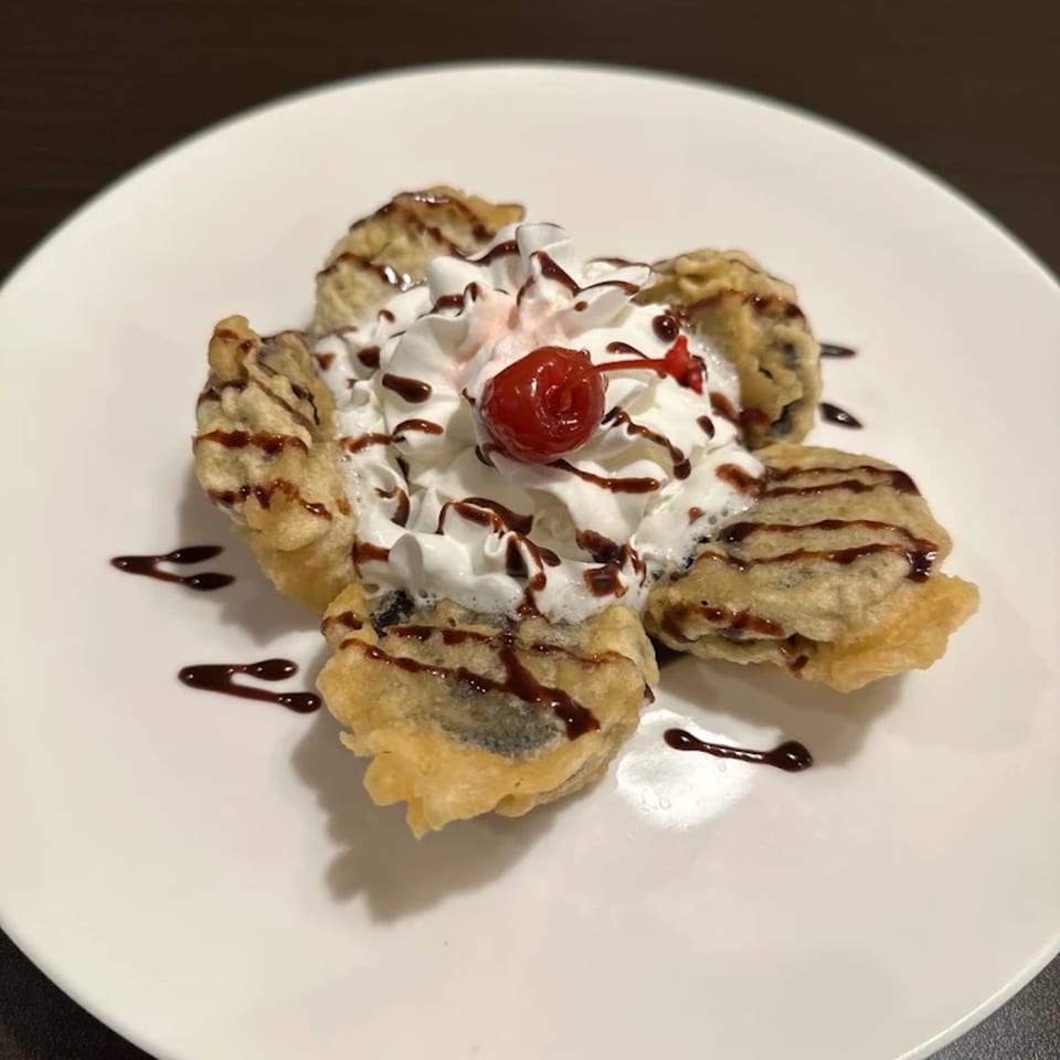 Deep-fried dessert with whipped cream, chocolate drizzle, and a cherry on a white plate.