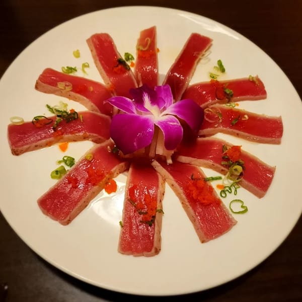 Seared tuna sashimi slices arranged in a circle with a purple orchid, tobiko, and scallions.