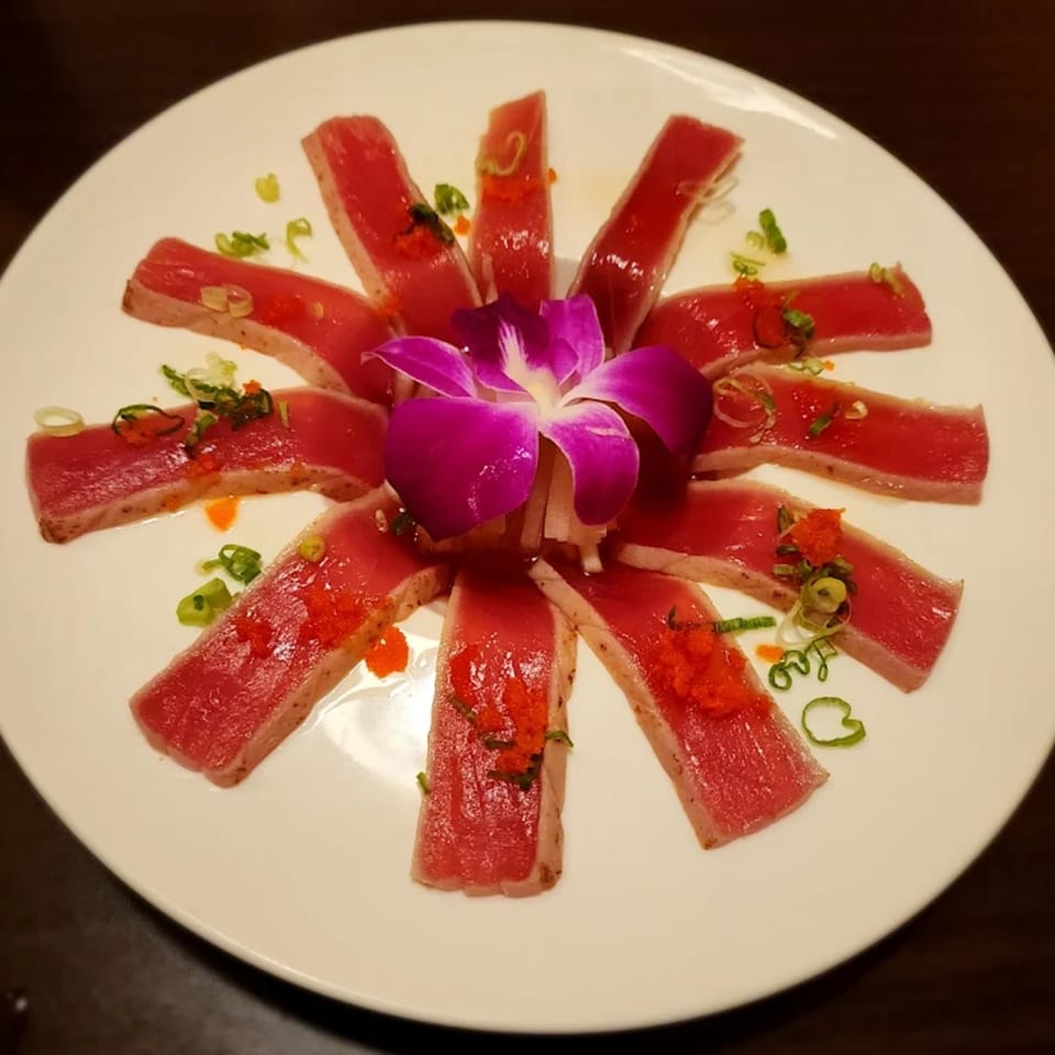 Seared tuna sashimi slices arranged in a circle with a purple orchid, tobiko, and scallions.