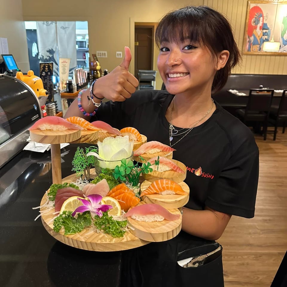 A smiling Flame Sushi staff member holds a large, tiered platter of sushi and sashimi.