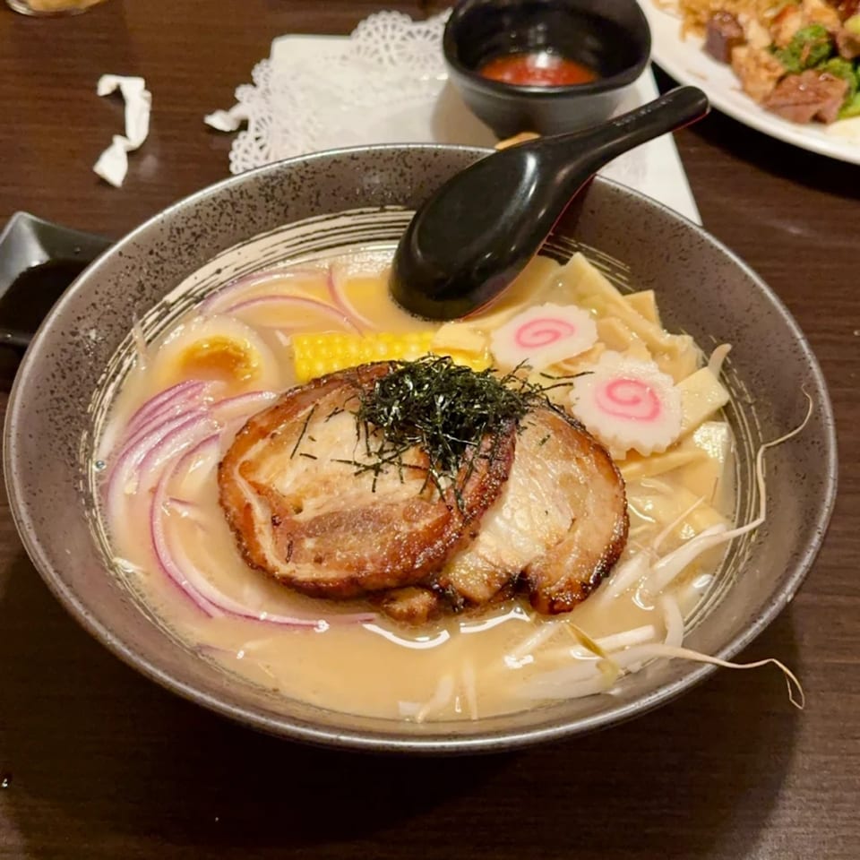 A close-up of a bowl of Flame Sushi ramen with pork, egg, corn, red onions, and fish cake.