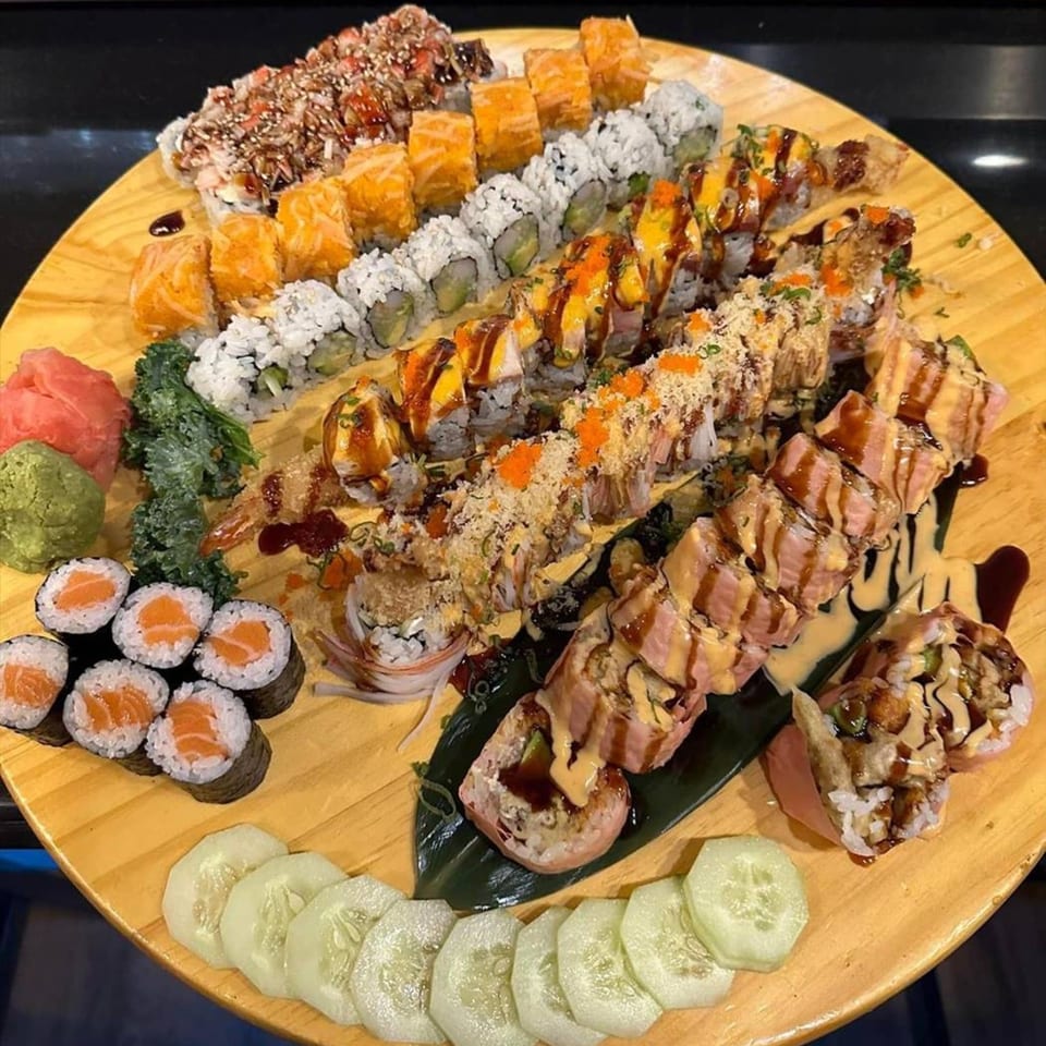 A large wooden platter filled with various Flame Sushi rolls, ginger, wasabi, and cucumber slices.